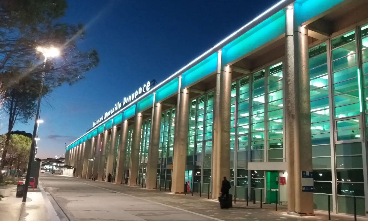Façade éclairée de l'aéroport Marseille-Provence à la tombée du soir, un voyageur rejoint le terminal avec sa valise à roulettes.