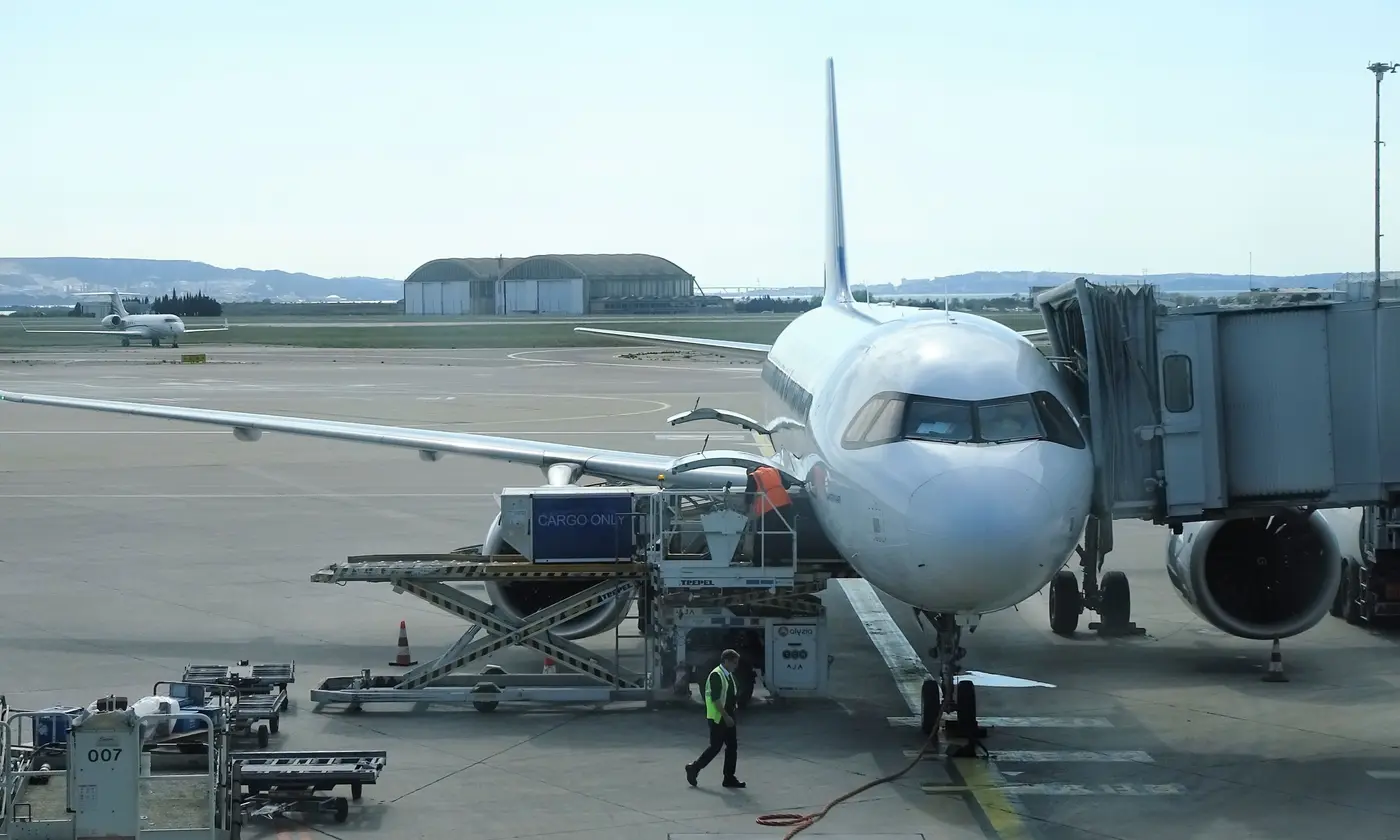 Avion de ligne stationné au contact à l'aéroport Marseille-Provence en plein jour, passerelle d'embarquement branchée, chargement des bagages en soute et agent de piste en gilet fluo.