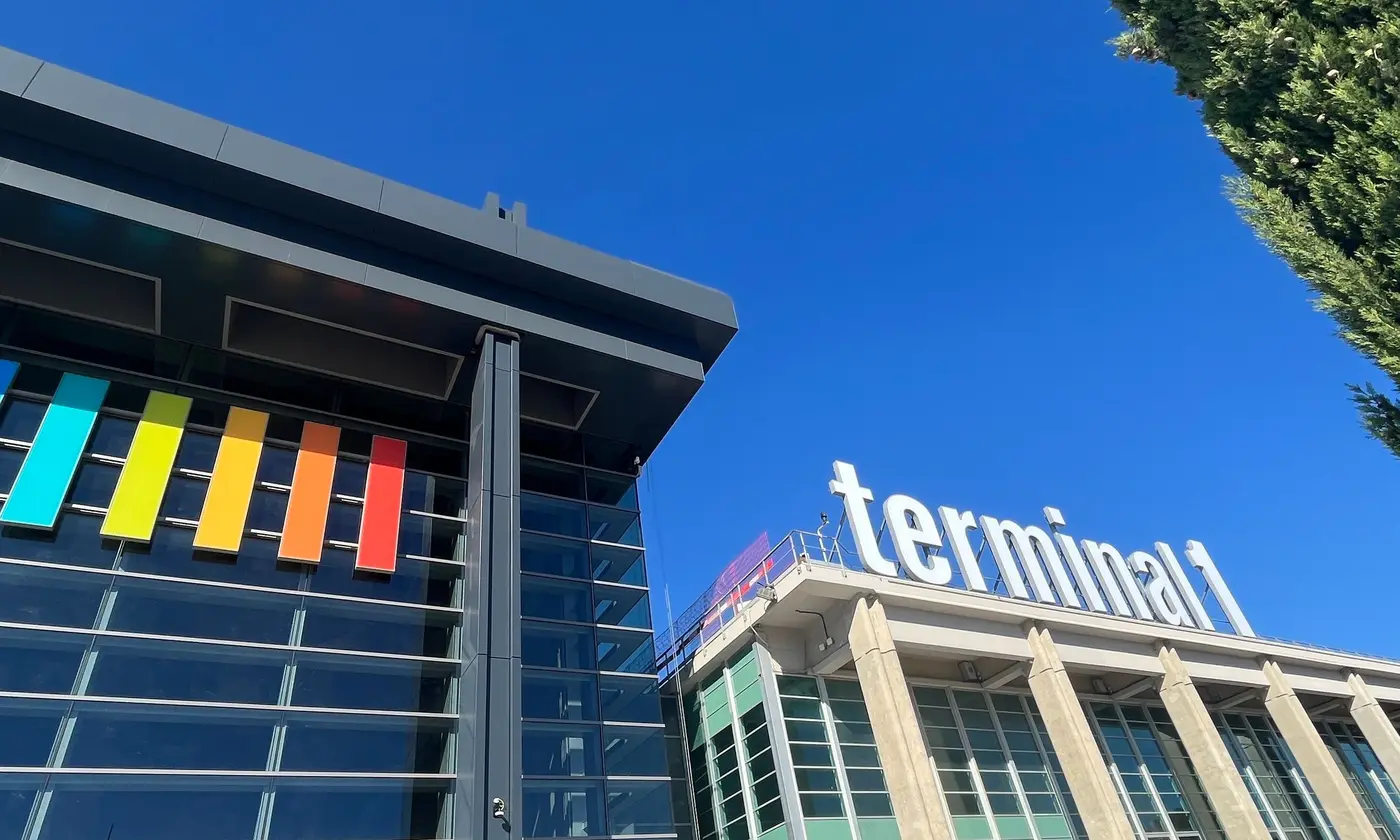 L'enseigne « terminal 1 » de l'aéroport Marseille-Provence et la façade de verre aux panneaux colorés du nouveau terminal, sous un ciel bleu de plein midi.
