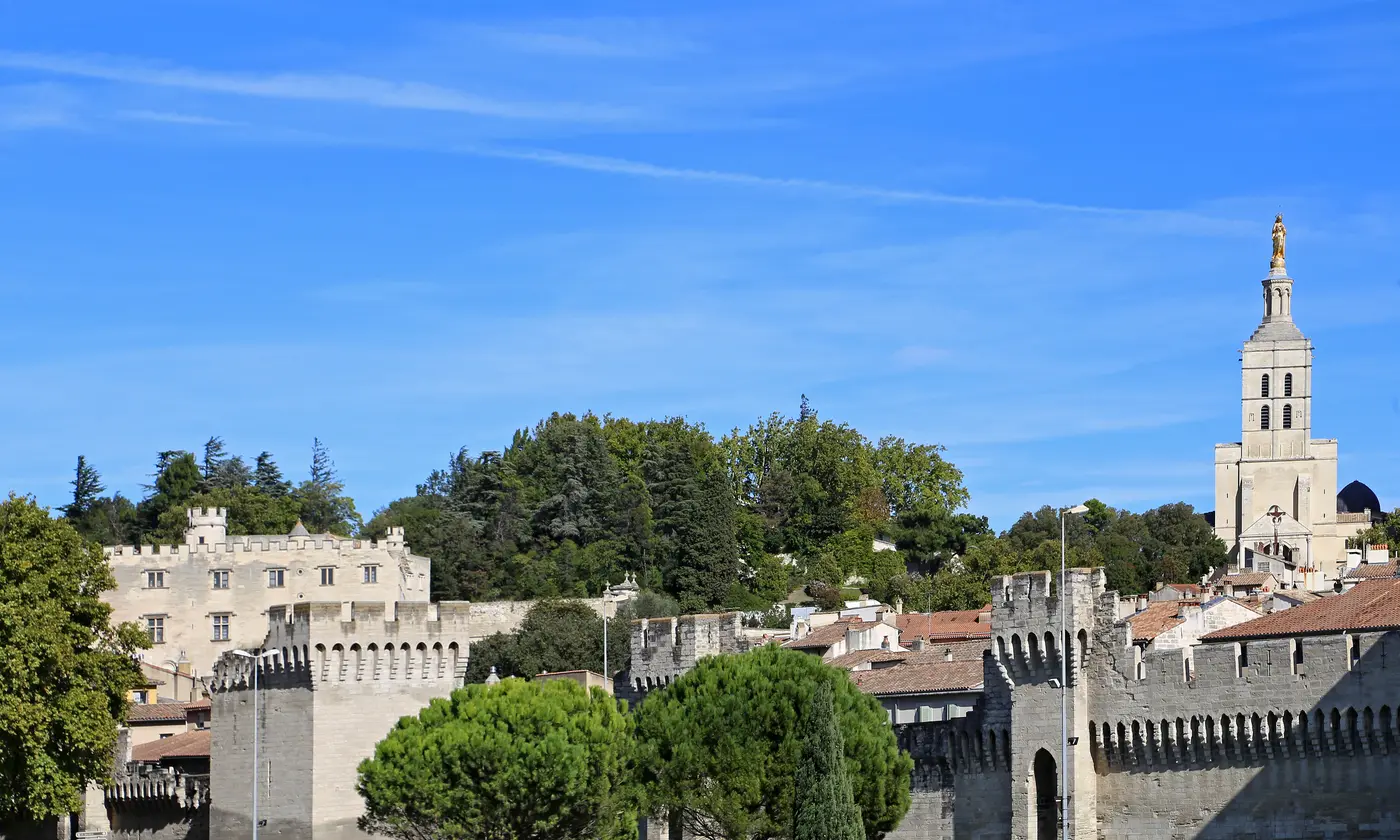 Les remparts médiévaux et leurs tours crénelées sous le ciel bleu, dominés par le clocher et la Vierge dorée de Notre-Dame des Doms, Avignon