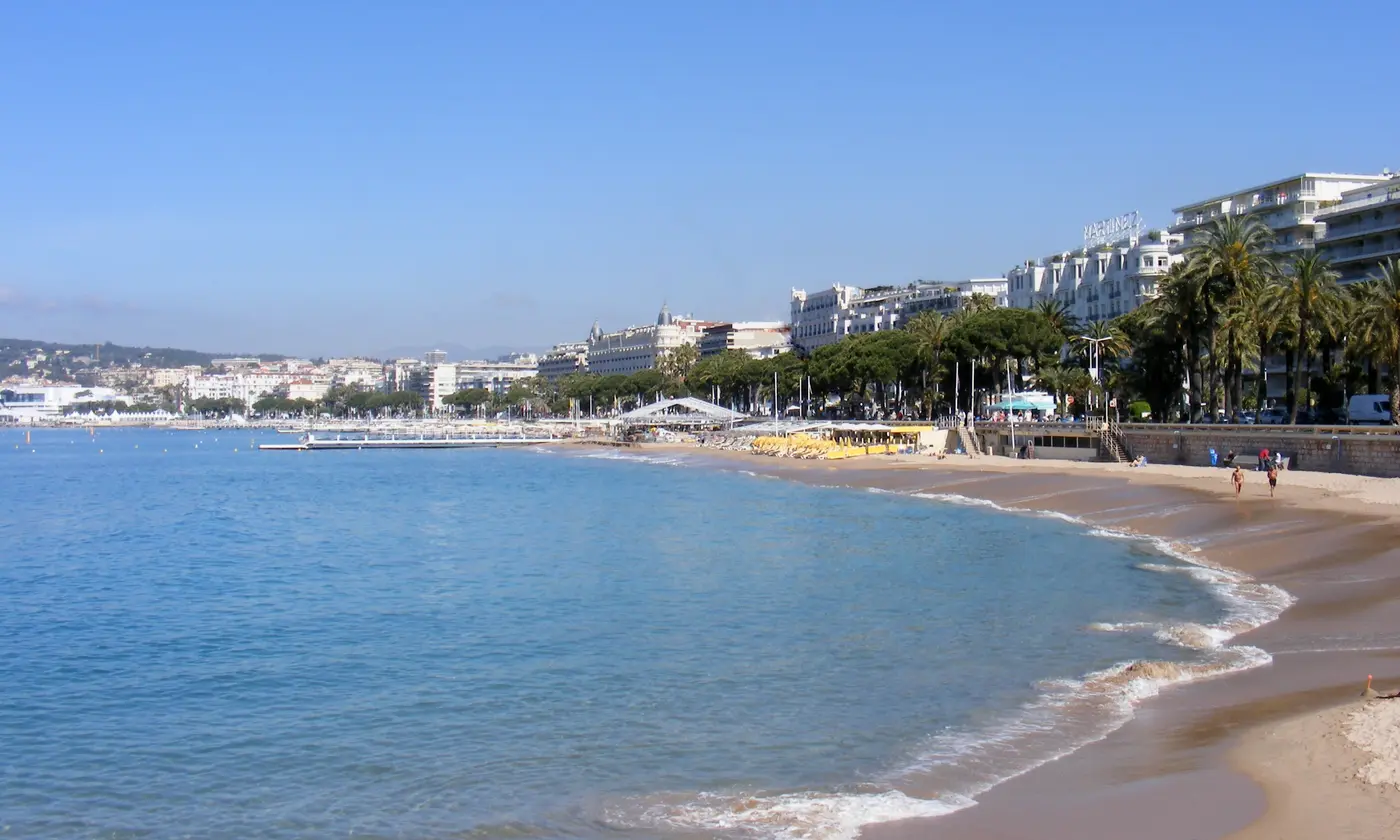 La courbe de sable de la plage de la Croisette, bordée de palmiers et de palaces, avec l'eau turquoise au premier plan, Cannes