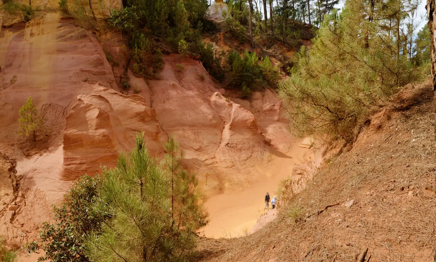 Falaises d'ocre orangées du Sentier des Ocres à Roussillon, pins et deux promeneurs minuscules sur le sable doré