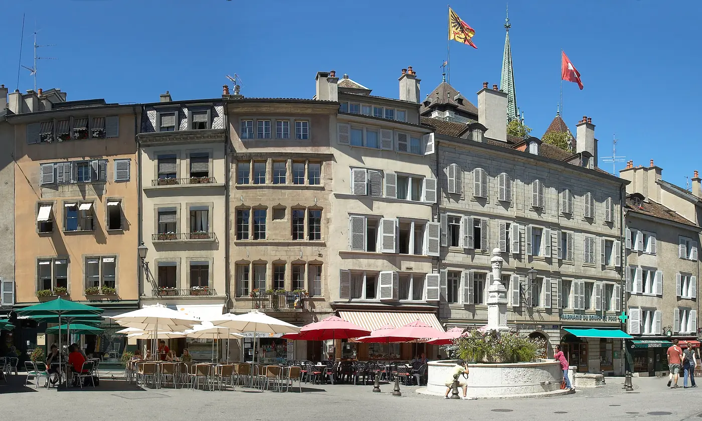 Façades, terrasses et fontaine de la place du Bourg-de-Four au cœur de la vieille ville, avec la flèche de la cathédrale Saint-Pierre en arrière-plan, Genève