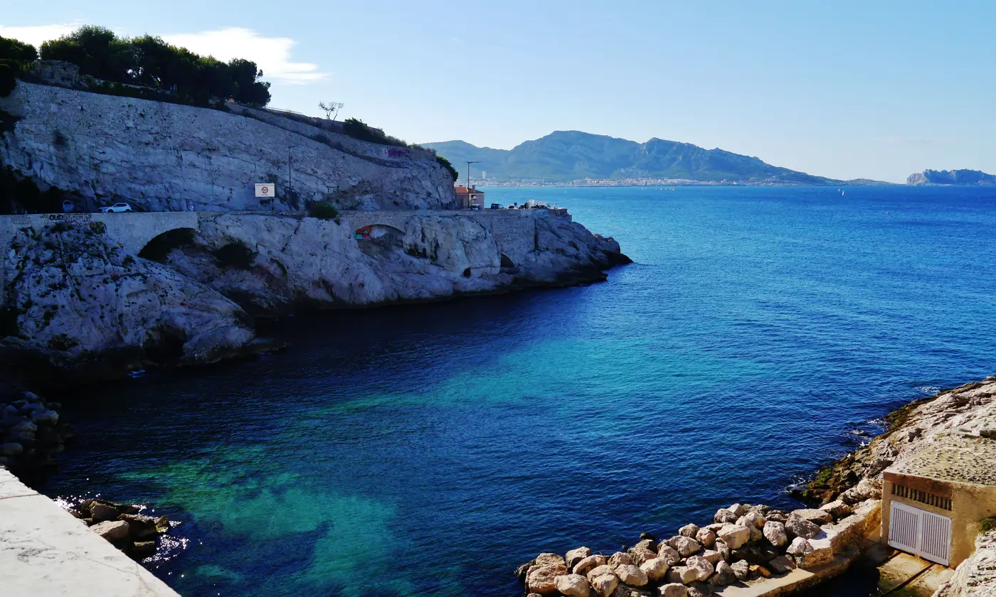 Crique aux eaux turquoise sous la Corniche Kennedy, avec les collines de Marseilleveyre à l'horizon.