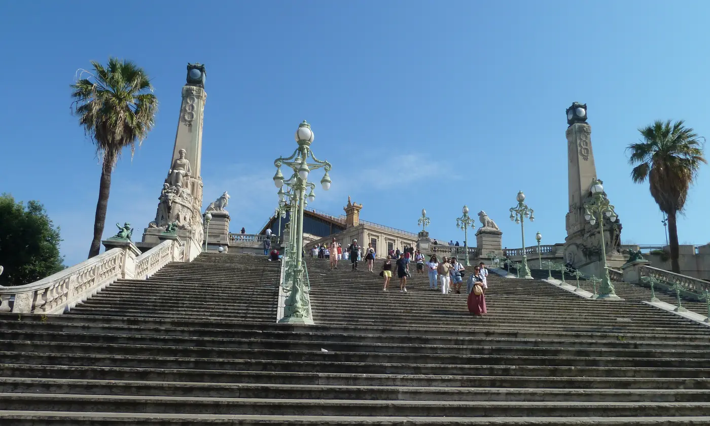 L'escalier monumental de la gare Saint-Charles, ses obelisques sculptes, ses lampadaires en fonte et ses palmiers sous un ciel d'ete.