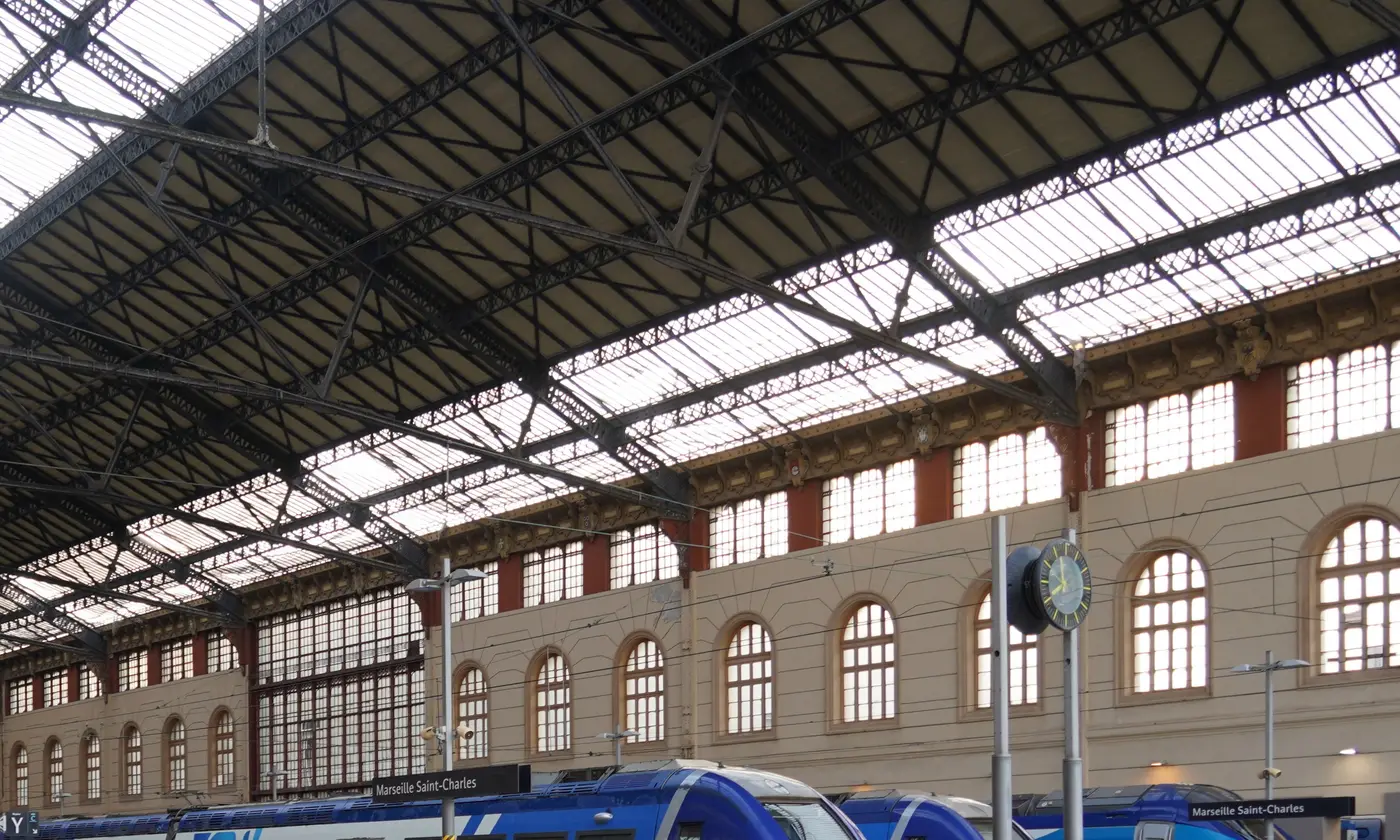 La charpente metallique et la verriere de la grande halle de Saint-Charles, l'horloge et la facade en pierre a arcades, au-dessus des trains regionaux a quai.