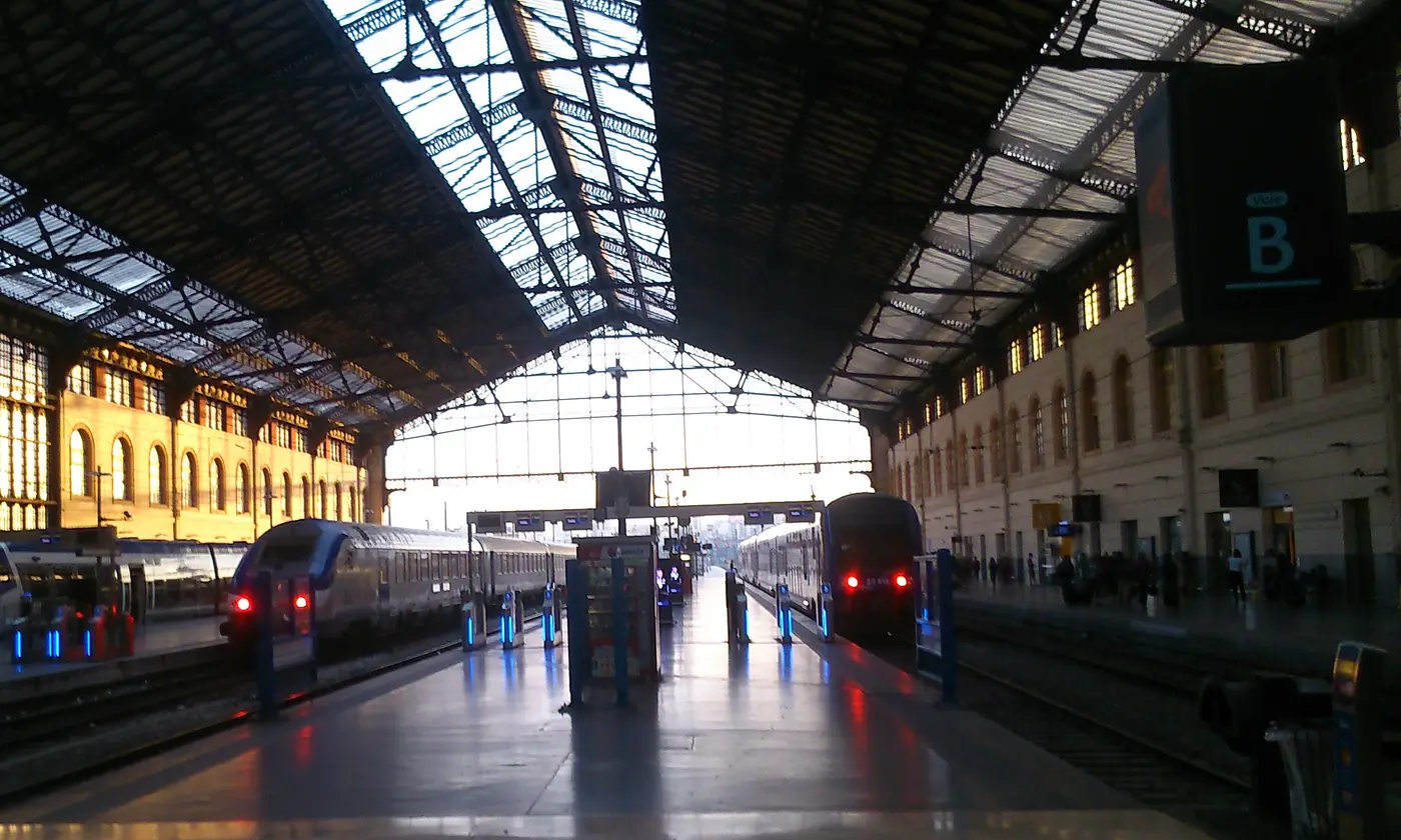 L'interieur de la grande halle de la gare Saint-Charles au petit matin : la verriere, la facade doree par le soleil levant, la signaletique Voie B et les trains a quai.
